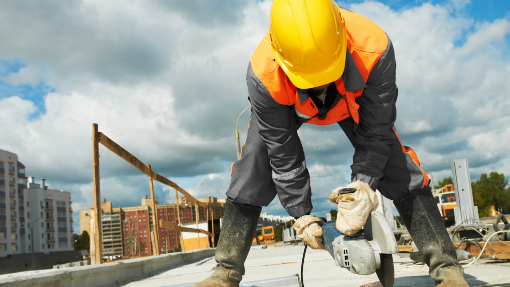 A hard-hat donning worker in a high-visibility jacket is seen. He is hunched over and using an angle grinder to saw through concrete.