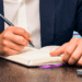 A formally dressed solicitor hunches over a notebook, pen in hand, taking client notes that will later be spun out into a comprehensive Will.