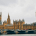 The golden Palace of Westminster, meeting place of the Houses of Parliament, is seen from across the Southbank across the river Thames. The sky is overcast and grey, contrasting with the bright radiance of Westminster itself.