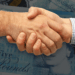 Two hands reach out, clasping each other in a firm but welcoming grasp. Behind this scene of relatable agreement, a lightly obscured stack of £5 sterling notes can be seen.