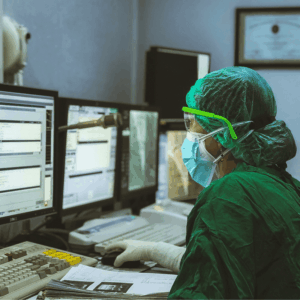 A medical professional dressed in full-body PPE reviews medical records on a desktop computer.