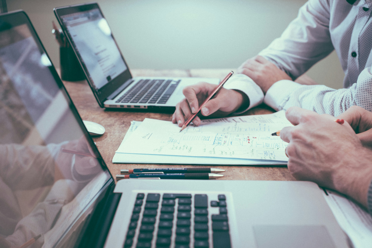 Two pairs of hands, both attached to formally dressed arms, are seen pointing to specific sections of a written document resting on a tabletop. Laptops are also seen open on the table, and we are left to infer that this is a negotiation of some description.